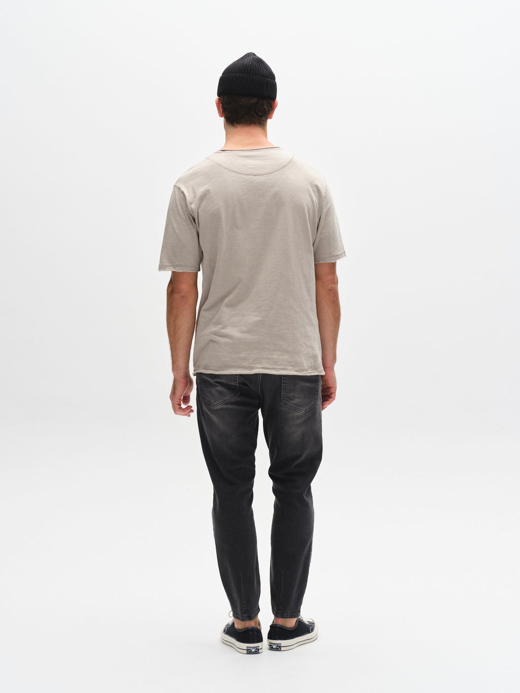 A person faces away from the camera, wearing a black beanie, dark jeans, slip-on shoes, and a Gabba GABkostant New SS - Grey Paloma t-shirt against a plain white background.