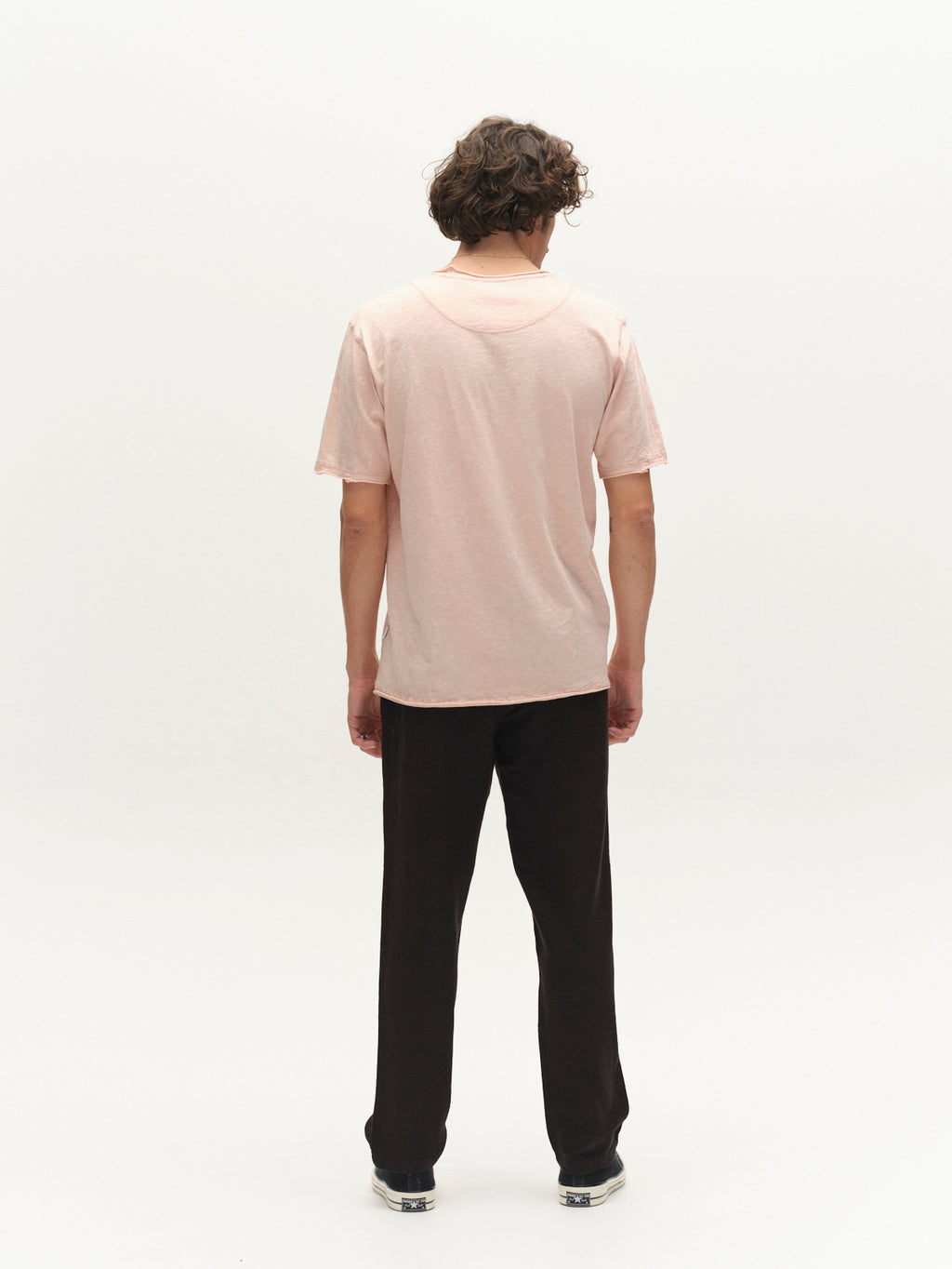 A person with curly hair faces away, wearing the Gabba GABkostant New SS t-shirt in Misty Rose, paired with black pants and dark sneakers, standing against a plain white background.
