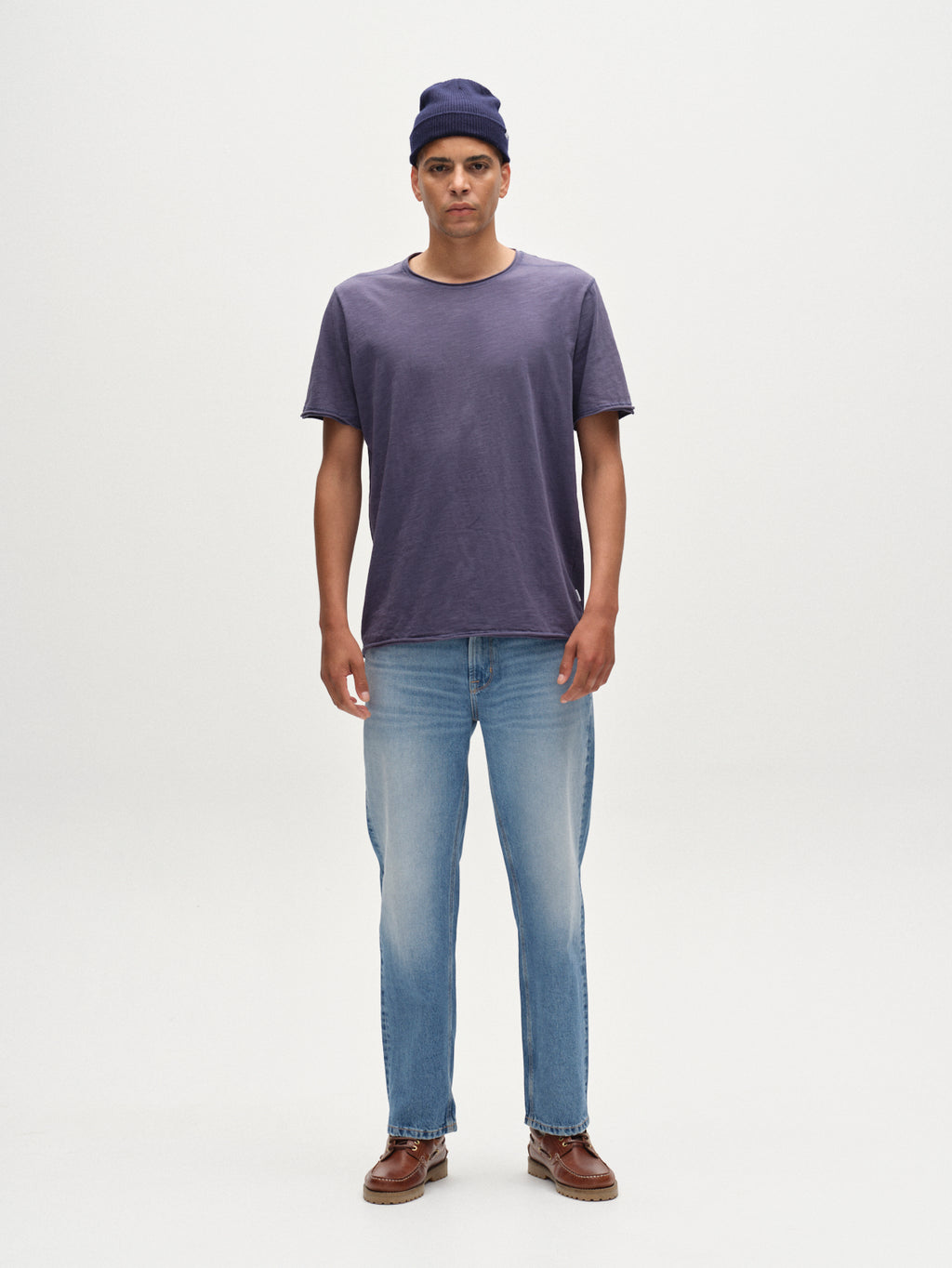 A man stands facing forward against a plain white background, wearing the Gabba Kostant New SS Vintage Indigo T-shirt in regular fit, paired with a blue beanie, light blue jeans, and brown shoes.