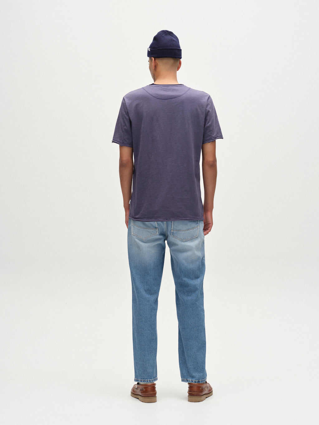 A person faces away from the camera wearing the Gabba Kostant New SS - Vintage Indigo t-shirt with light blue jeans, brown shoes, and a navy beanie, posed against a plain white background.