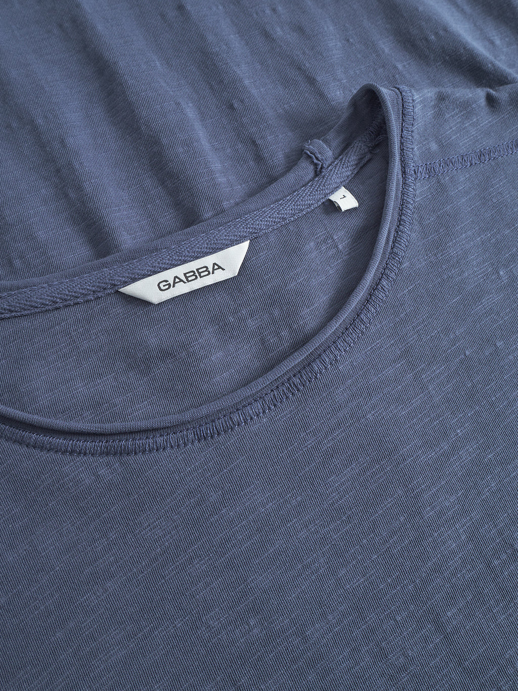 A close-up of the neckline of Gabbas Kostant New SS T-shirt in Vintage Indigo shows a white GABBA label inside the collar. The shirt is laid flat, highlighting its soft cotton slub fabric and regular fit.