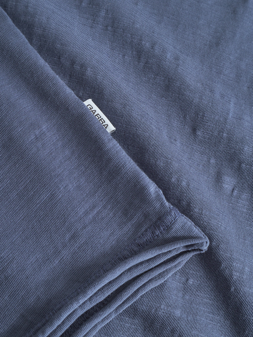 A close-up of folded Kostant New SS - Vintage Indigo fabric from Gabba, with a small white Gabba tag visible between the folds and clearly showing the cotton slub texture.