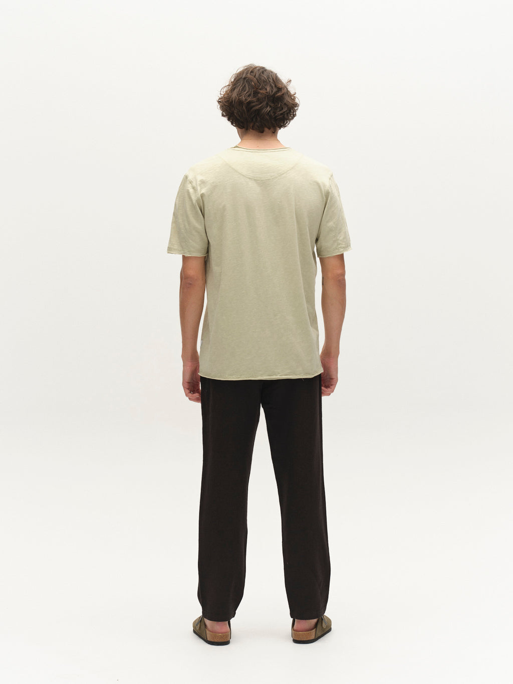 A person with short, curly hair faces away from the camera, wearing the Gabba GABkostant New SS t-shirt in Alfa Green, dark pants, and sandals against a plain white background.