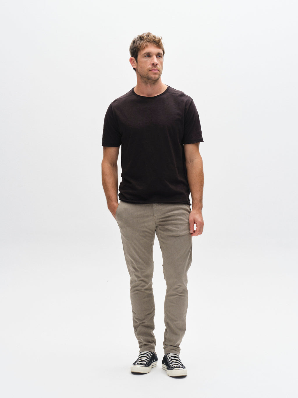 A man with short hair stands against a plain white background, wearing the Gabba GABkostant New SS - Meteorite black slub t-shirt, light tan pants, and black-and-white sneakers. One hand in his pocket adds an effortless, textured vibe to the outfit.