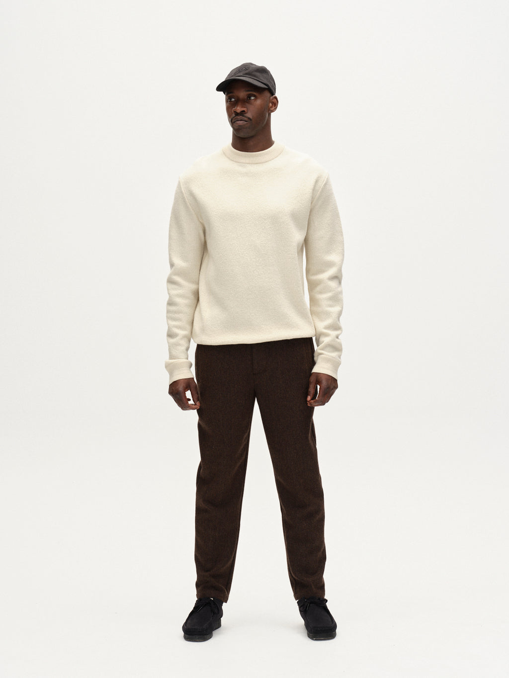 A man stands against a plain white background, wearing the Gabba Monza Wook - Brown tailored trousers with a tapered fit, a cream sweater, black shoes, and a black baseball cap. He faces forward with a neutral expression.
