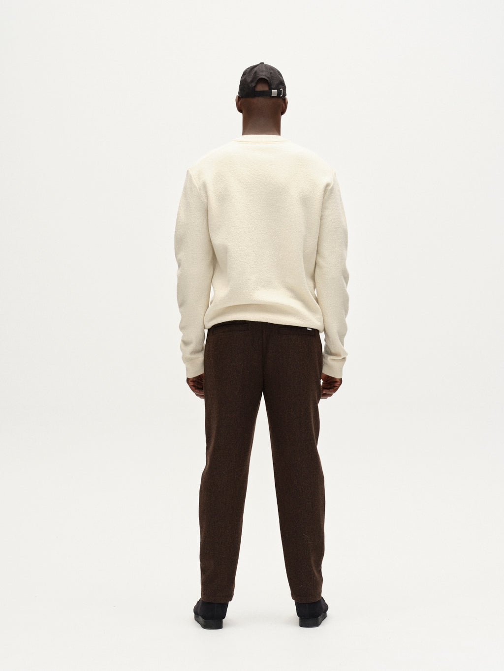 A person faces away from the camera, wearing Gabba’s Monza Wook - Brown tailored trousers with a tapered fit, a cream sweater, black cap, and black shoes against a plain white background.