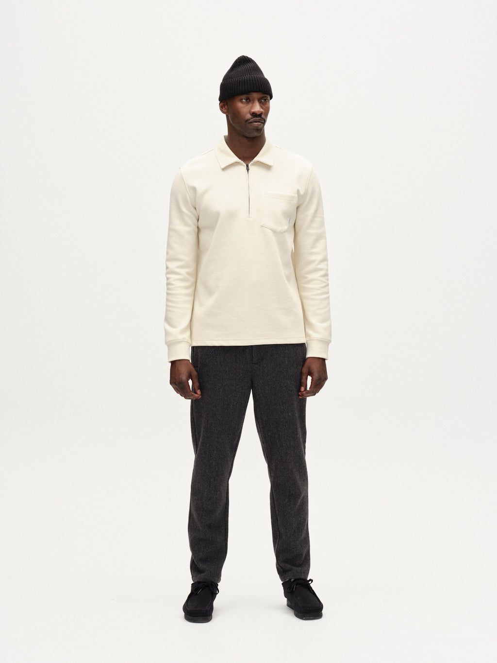 A man stands against a plain white background, looking slightly to his right, wearing a cream long-sleeve polo shirt, Gabba Monza Wook trousers in dark grey with a regular tapered fit, black shoes, and a black knit beanie.