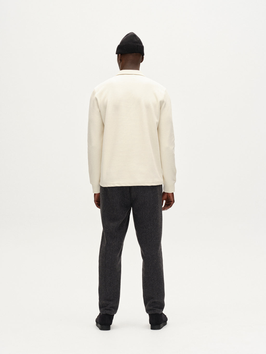 A person faces away from the camera against a plain white background, wearing Gabba Monza Wook - Dk. Grey tailored trousers with a regular tapered fit, a cream long-sleeve shirt, black shoes, and a black beanie.