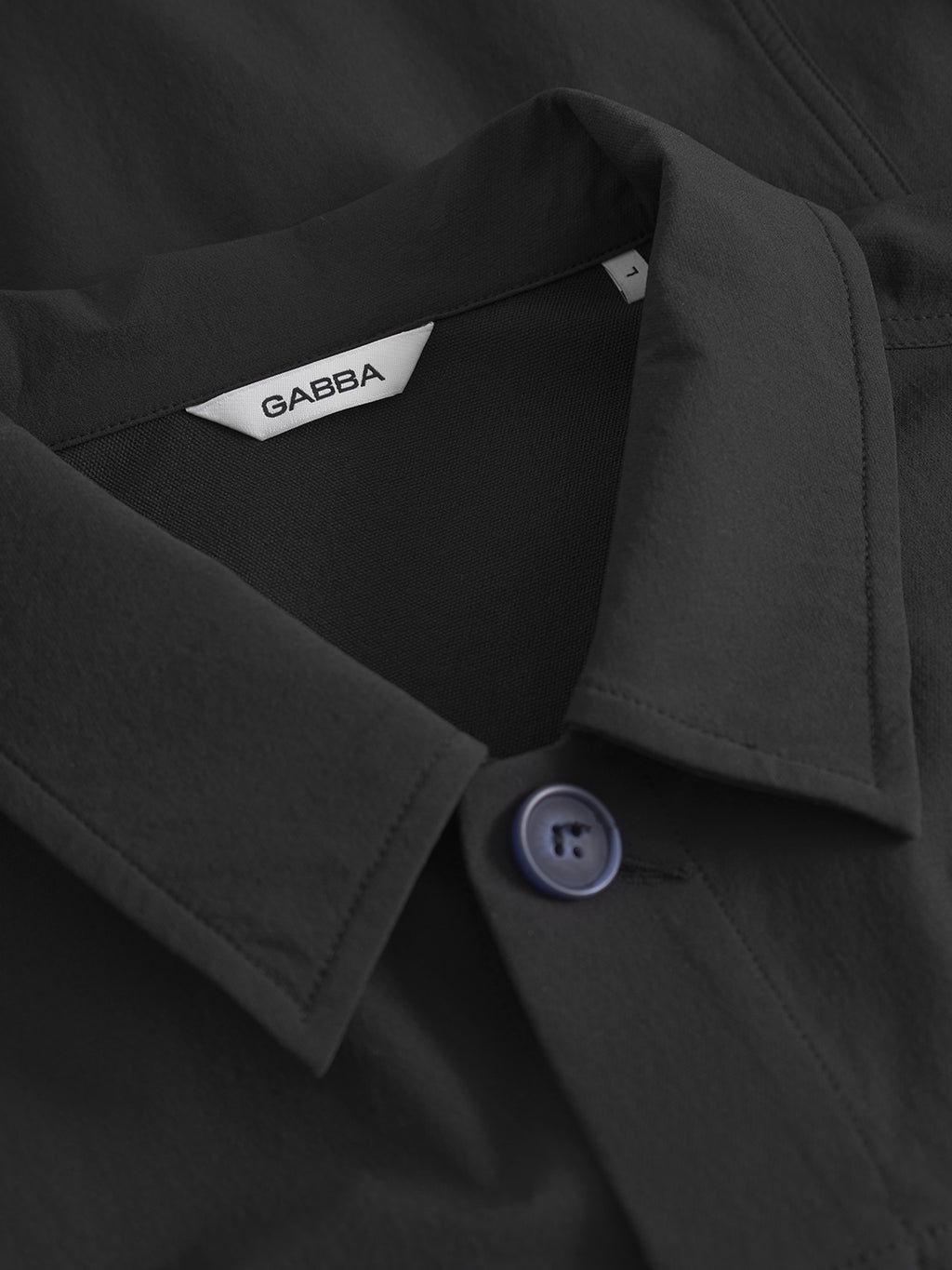 Close-up of the Gabba Koy Tech - Black shirt featuring a blue button, a visible white Gabba label inside the collar, and smooth stretch tech fabric for a neatly tailored look.