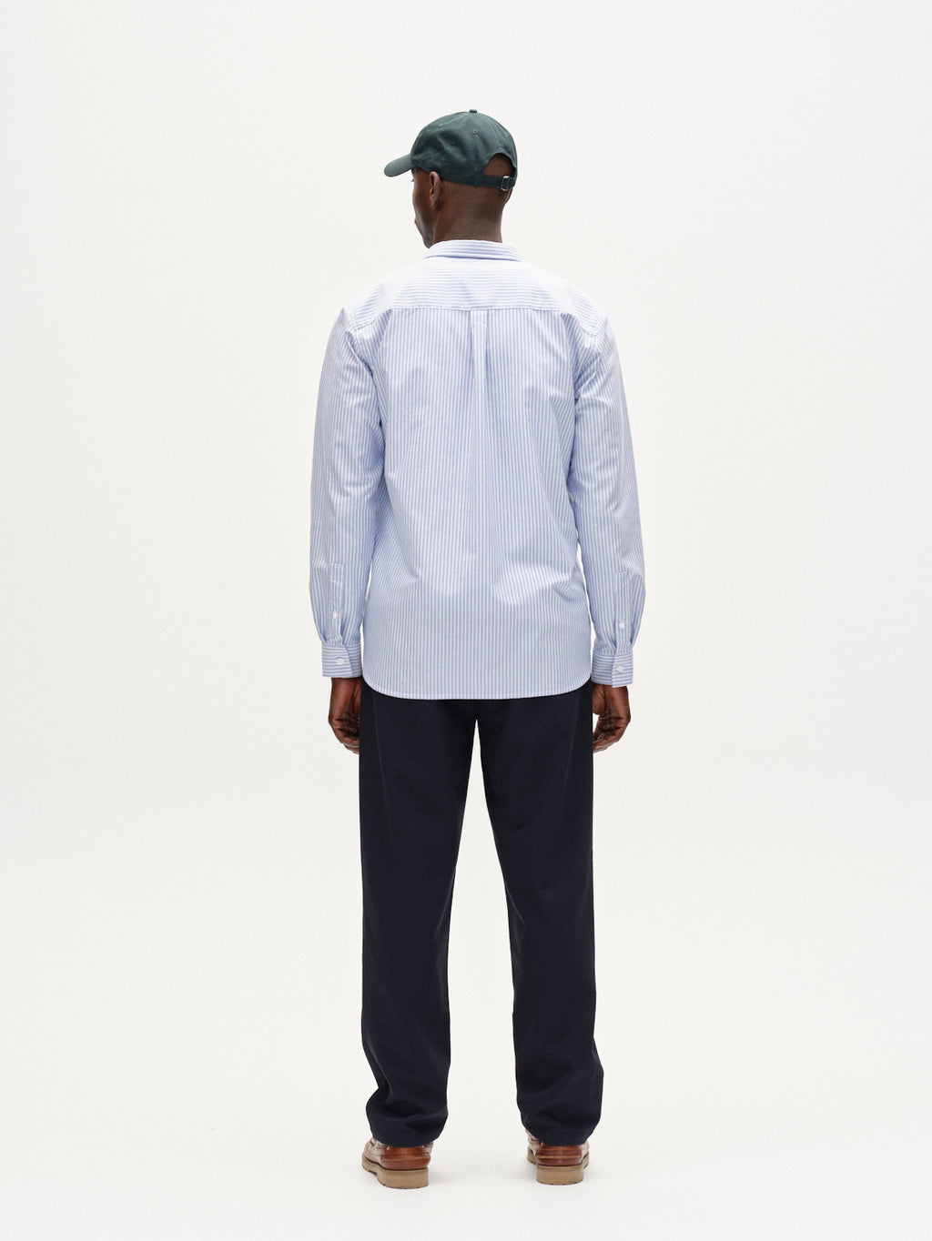 A man faces away against a plain white background, wearing Gabbas Napoli 19 Phin GRS - Navy tailored pants with a dark green baseball cap, light blue and white striped button-up shirt, and brown shoes.