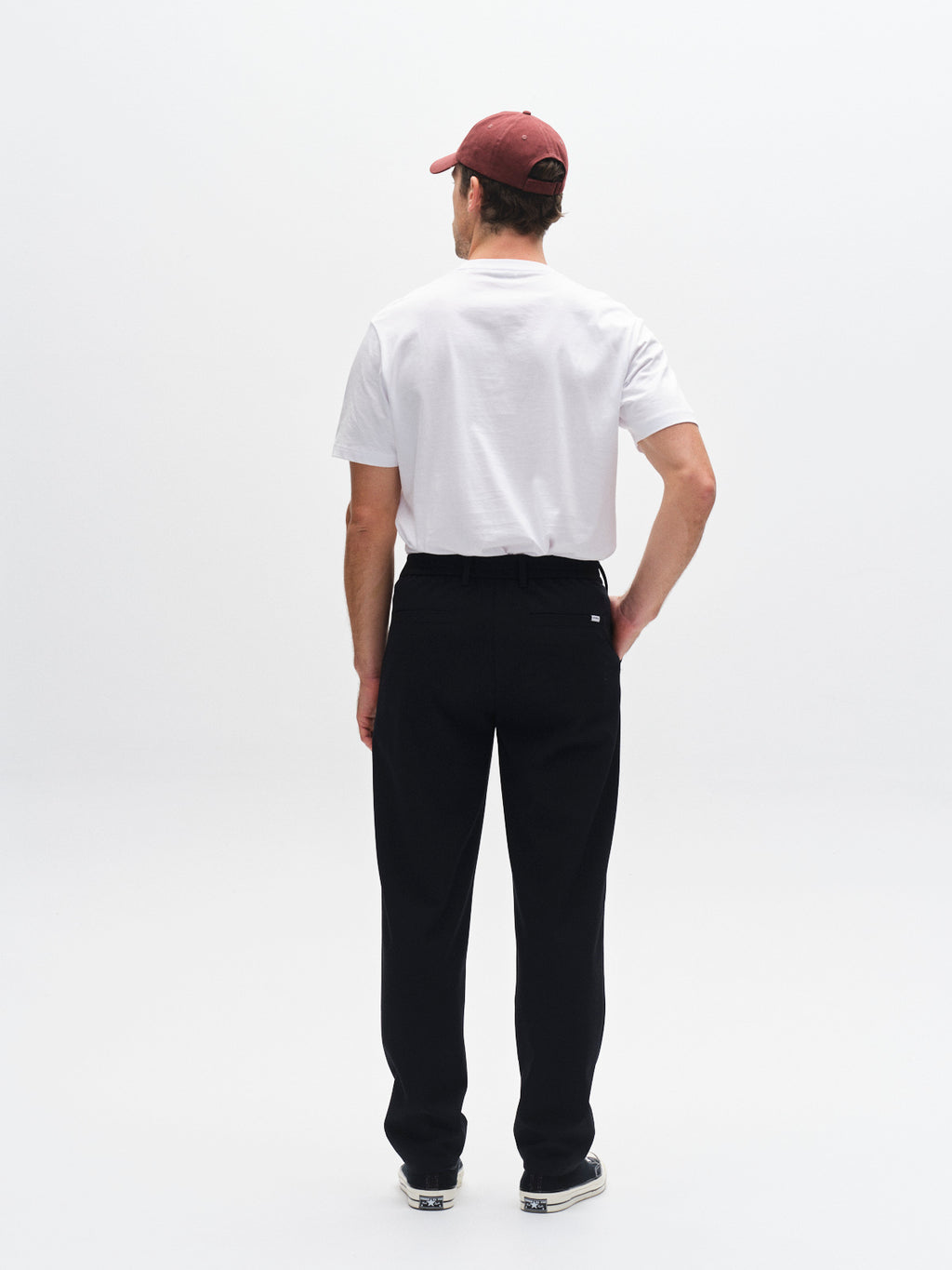 A man stands facing away from the camera, wearing a white T-shirt, black and white sneakers, a maroon baseball cap, and Gabba GABnapoli 19 Phin GRS - Blue Black pants against a plain white background.