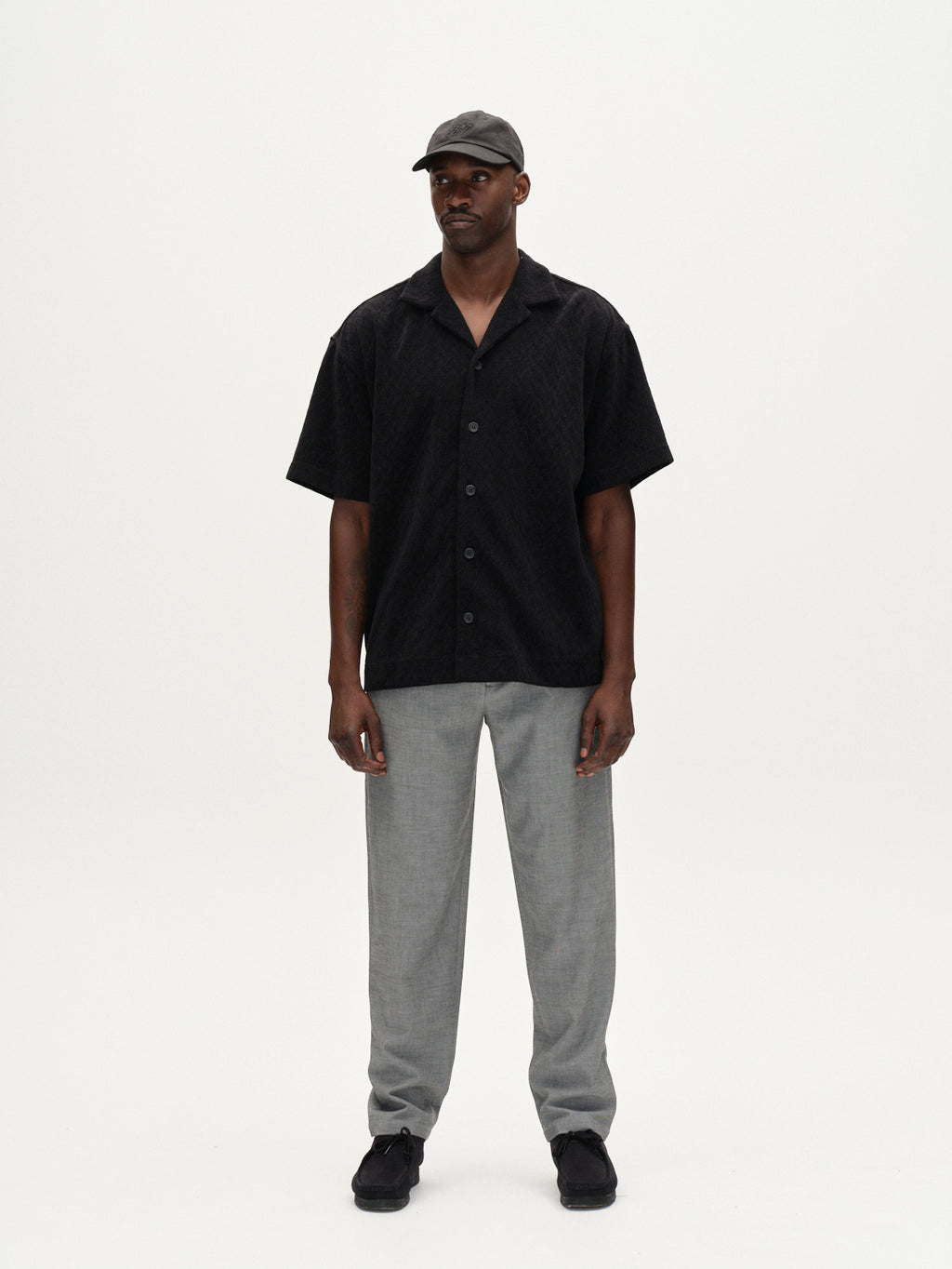 A man stands against a plain white background in classic menswear: a black short-sleeve button-up, Gabba Napoli 19 Phin GRS - Lt. Grey trousers, black shoes, and a dark baseball cap. He faces forward with a neutral expression.