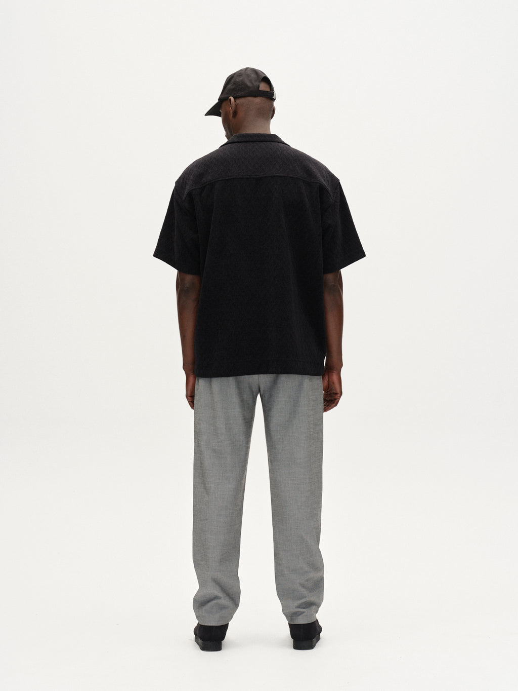 A person stands facing away against a plain white background, wearing Gabba Napoli 19 Phin GRS - Lt. Grey trousers, a black short-sleeve shirt, black shoes, and a black cap.