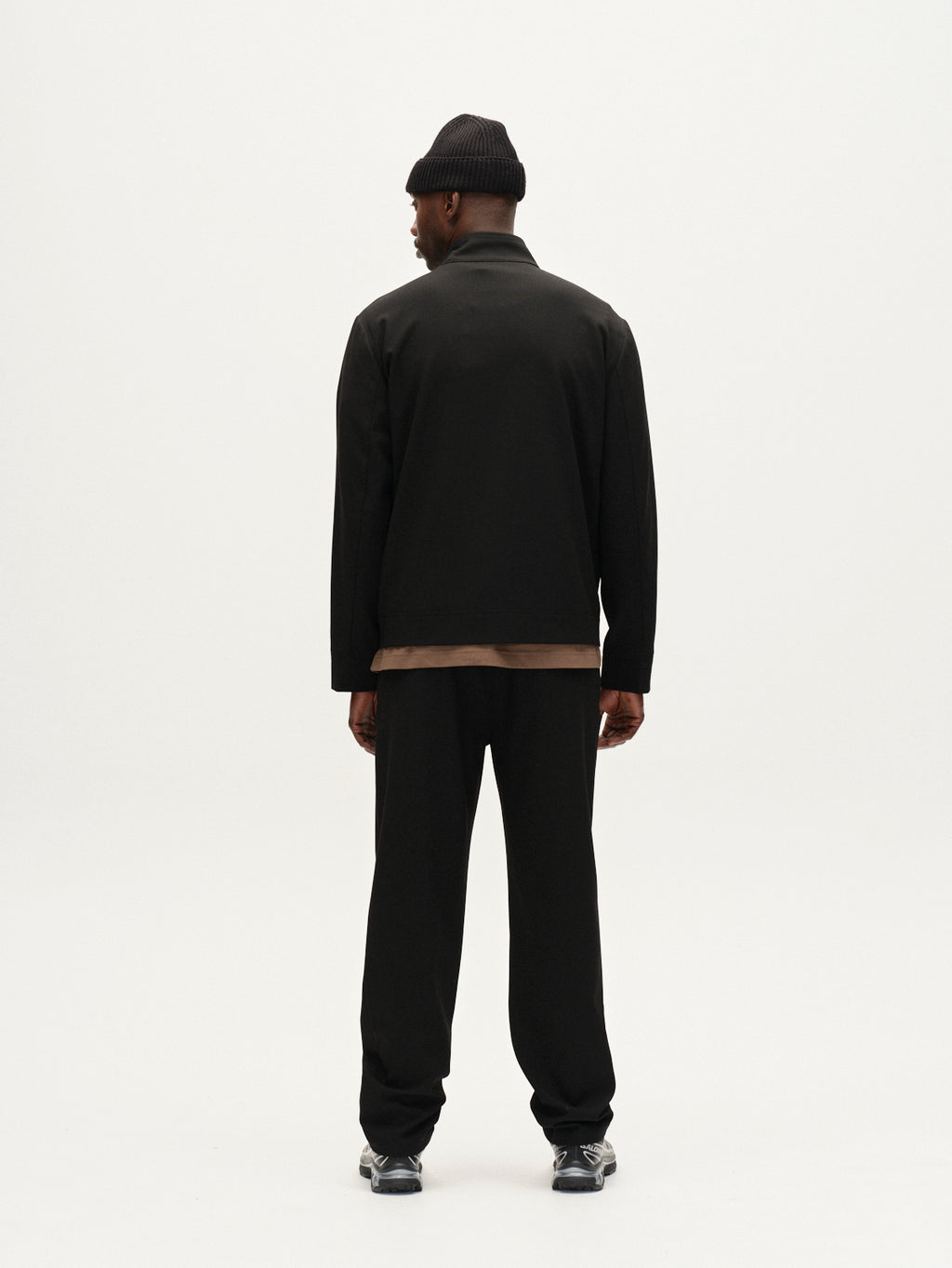 A man faces away from the camera, wearing Gabbas Napoli 19 Phin GRS - Black wool blend pleated trousers, a black beanie, black jacket over a brown shirt, and black sneakers against a plain white background.