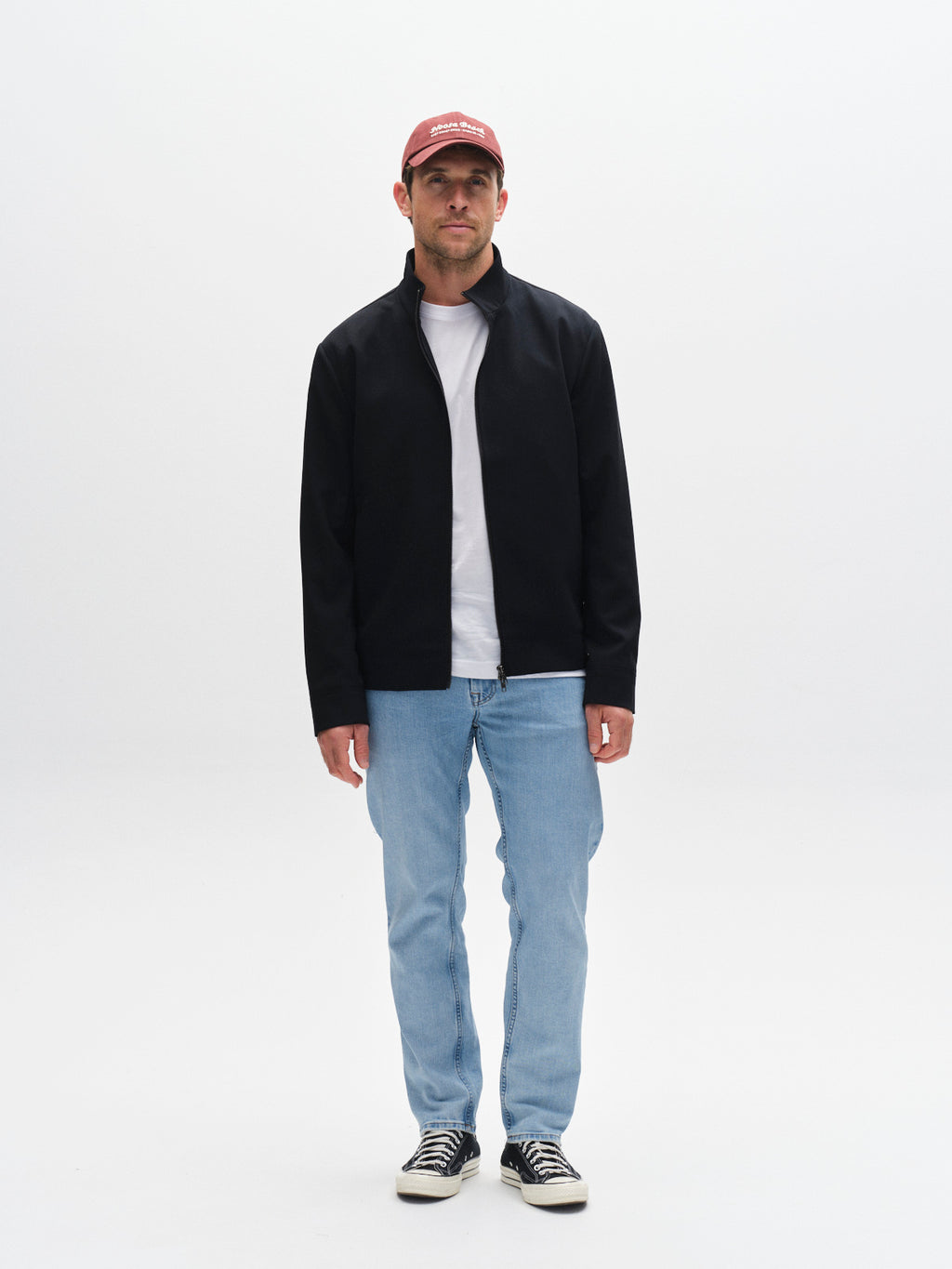 A man faces forward with a neutral expression against a plain white background, wearing the Gabba GABnik Phin GRS - Blue Black jeans, a red cap, a regular fit zip-through jacket, and black-and-white sneakers.