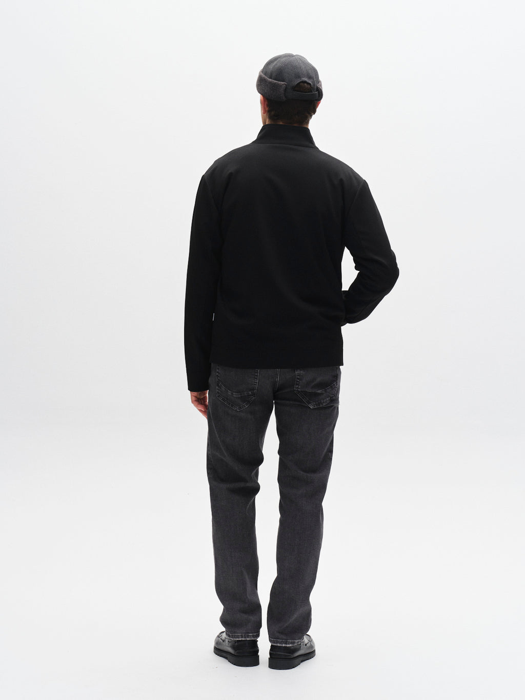 A person wearing the Gabba GABnik Phin GRS - Black zip-through jacket, dark jeans, black shoes, and a gray flat cap stands facing away in a bright, plain studio setting.