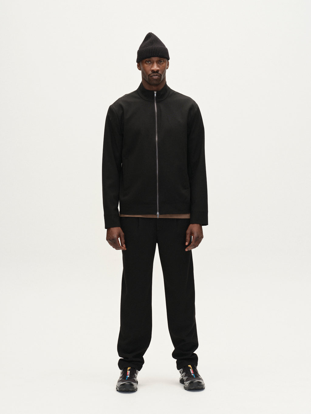 A man stands facing forward against a plain white background, wearing the Gabba Nik Phin GRS - Black zip-through jacket, paired with a black beanie, pants, and sneakers.