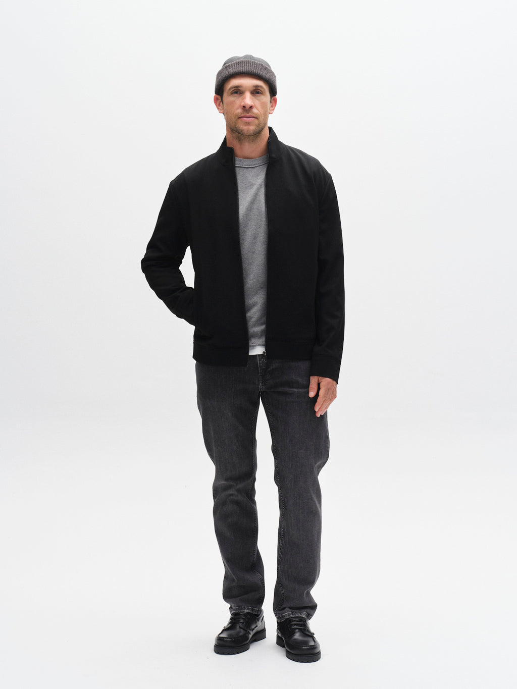 A man stands against a white background, wearing the Gabba GABnik Phin GRS - Black zip-through jacket over a gray shirt, paired with dark jeans, black shoes, and a gray beanie. One hand is in his pocket and he has a neutral expression.