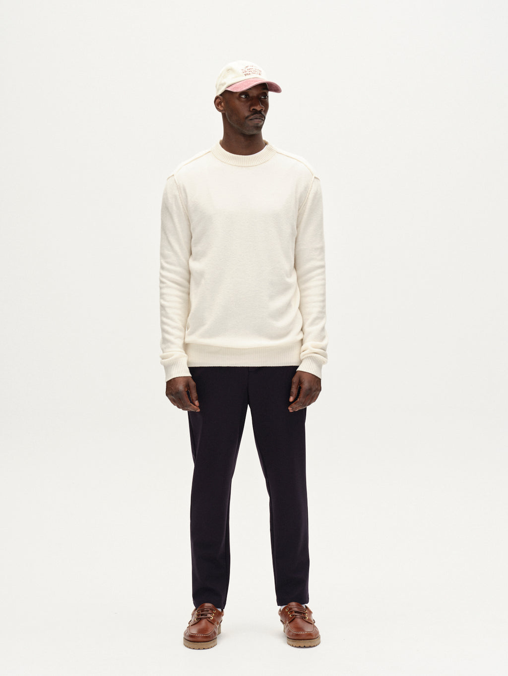 A man stands against a white background wearing a cream sweater, Monza Bascu - Navy straight fit wrinkle-resistant pants by Gabba, brown boat shoes, and a light baseball cap. He looks slightly left with his hands at his sides.