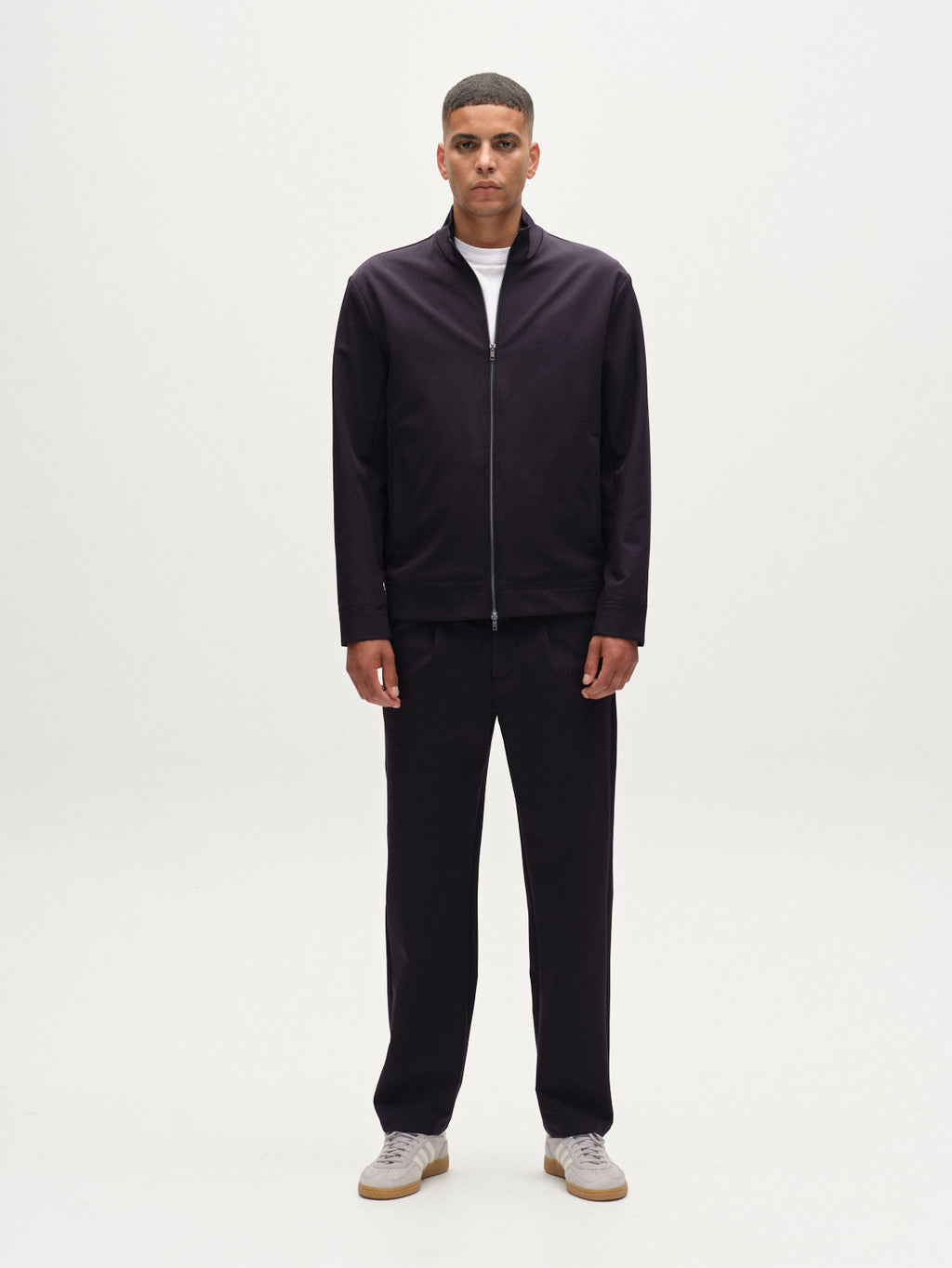 A man faces forward against a white background, wearing the Gabba Nik Bascu - Navy jacket over a white shirt, matching dark pants, and light gray sneakers.