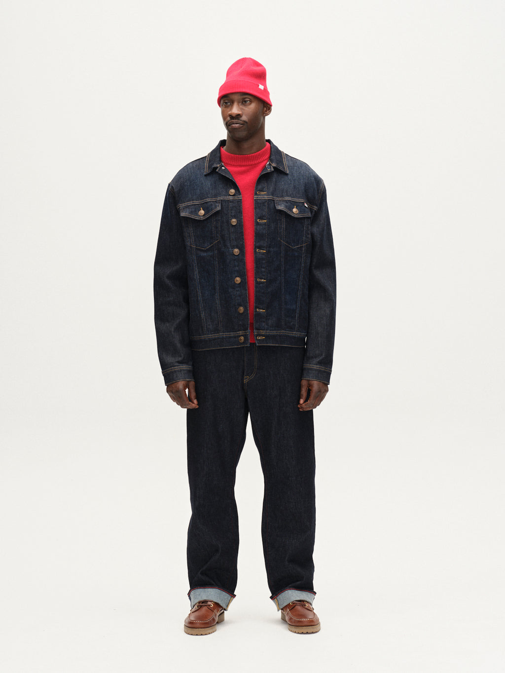 A man stands against a plain white background wearing a red beanie, a red shirt, a dark denim jacket, Gabba’s Noah Self - Dark Blue Denim loose straight jeans made from sustainable denim, and brown sandals. He looks slightly to the side.