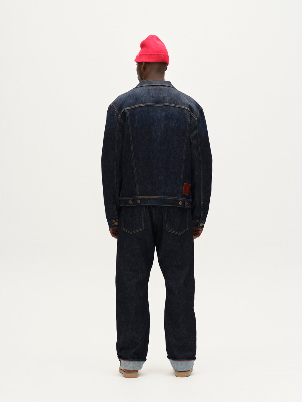 A person faces away from the camera, wearing Gabba’s Noah Self - Dark Blue Denim jacket and loose straight jeans made from recycled fibers, paired with a bright pink beanie and light brown shoes against a plain white background.