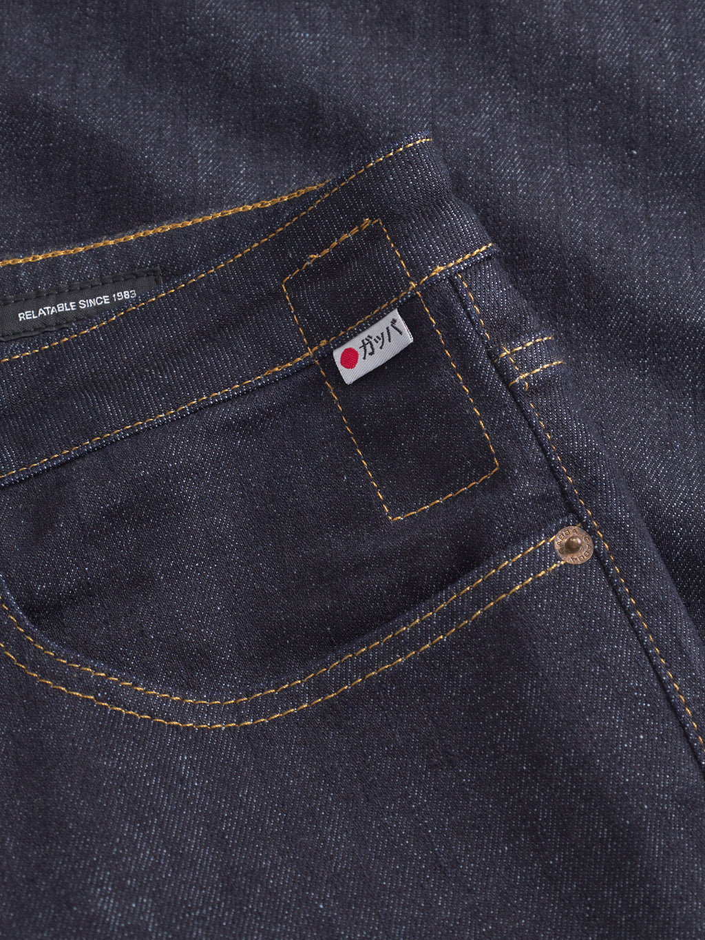 Close-up of Gabbas Noah Self - Dark Blue Denim jeans, featuring a front pocket with yellow stitching, a small white label with Japanese text and a red dot, and part of a metal rivet. RELATABLE SINCE 1982. is visible near the waistband.