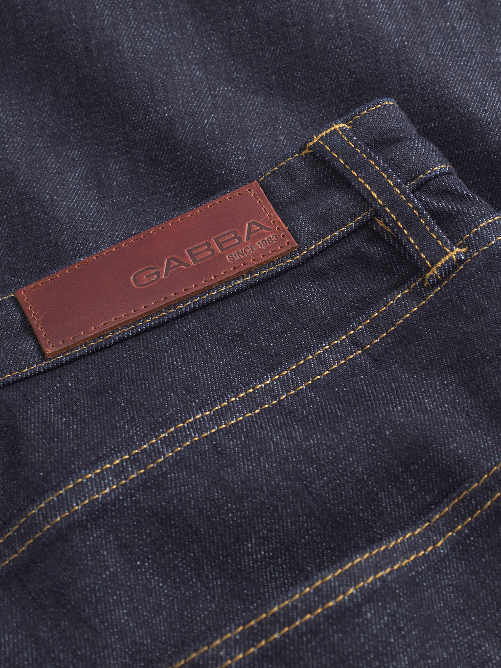 Close-up of Gabba Noah Self dark blue denim jeans with yellow stitching and a brown leather patch above the pocket, embossed with Gabba and Since 1983.