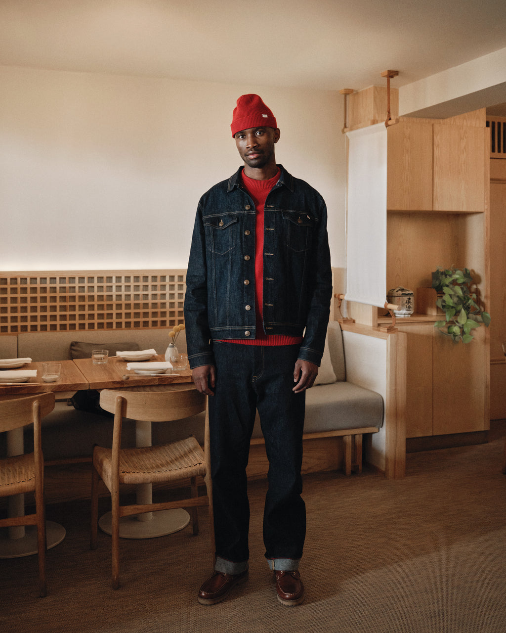 A man stands in a cozy, modern restaurant wearing a red beanie, red sweater, recycled denim jacket, brown shoes, and Gabba’s Noah Self - Dark Blue Denim jeans. Wooden furniture and neutral décor complete the background.