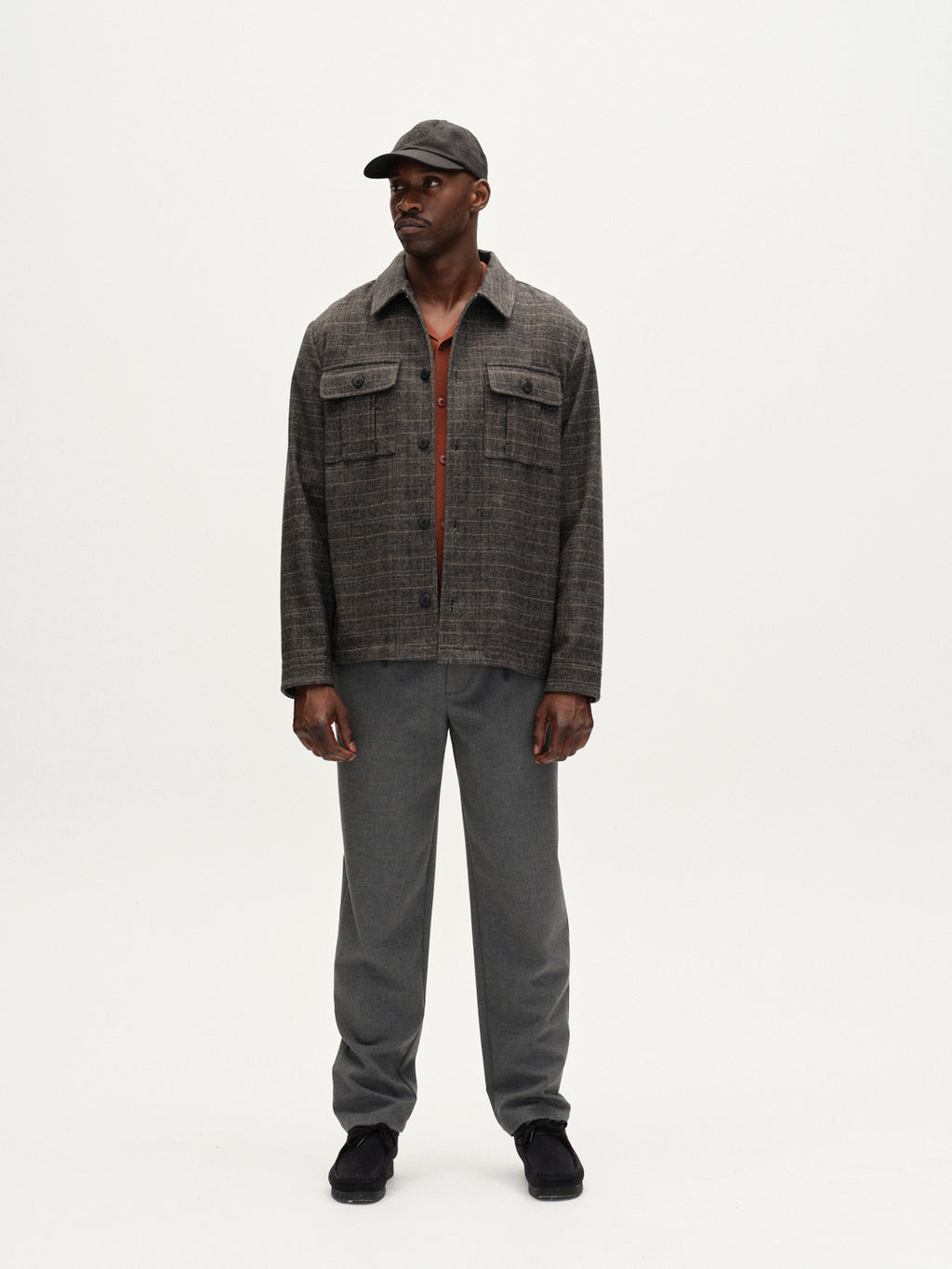A man stands against a white background, wearing Gabba’s Flap Tishi in Brown Check over a brown shirt, paired with gray pants, black shoes, and a dark cap. He faces forward with his hands at his sides.