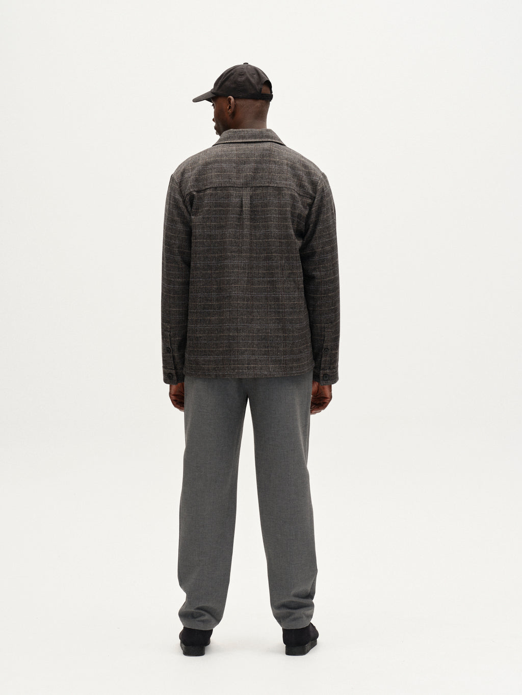 A man faces away from the camera, wearing the Gabba Flap Tishi overshirt in brown check, paired with gray pants, black shoes, and a black baseball cap, set against a plain white background.