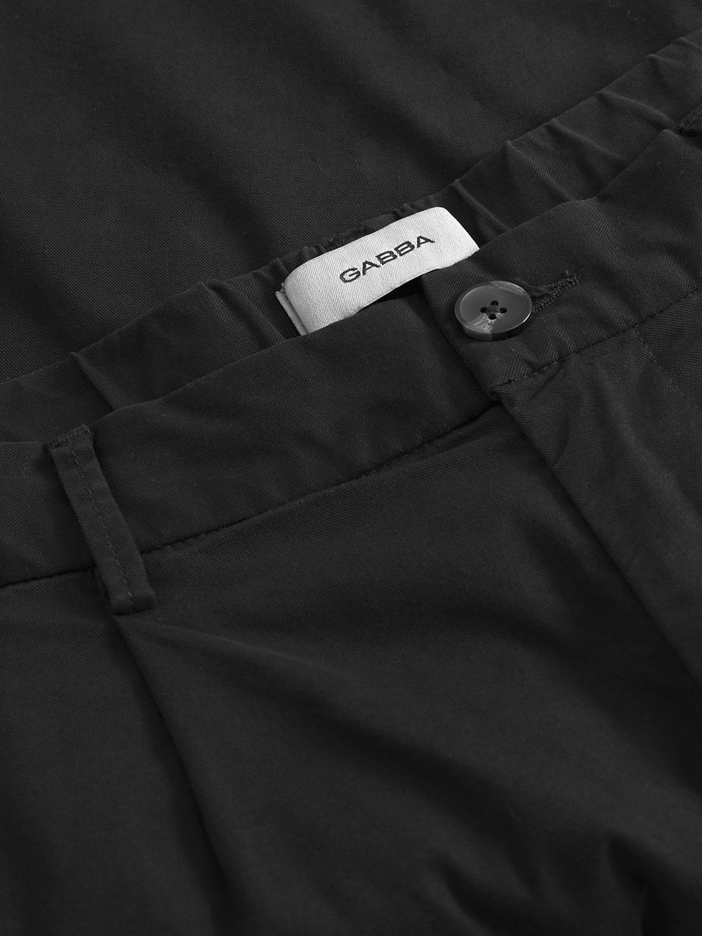 Close-up of the waistband of Imola Kuadro - Black wide-leg pants with a button and a white Gabba label. Crafted from smooth, garment-dyed stretch fabric, these pants feature neat stitching.