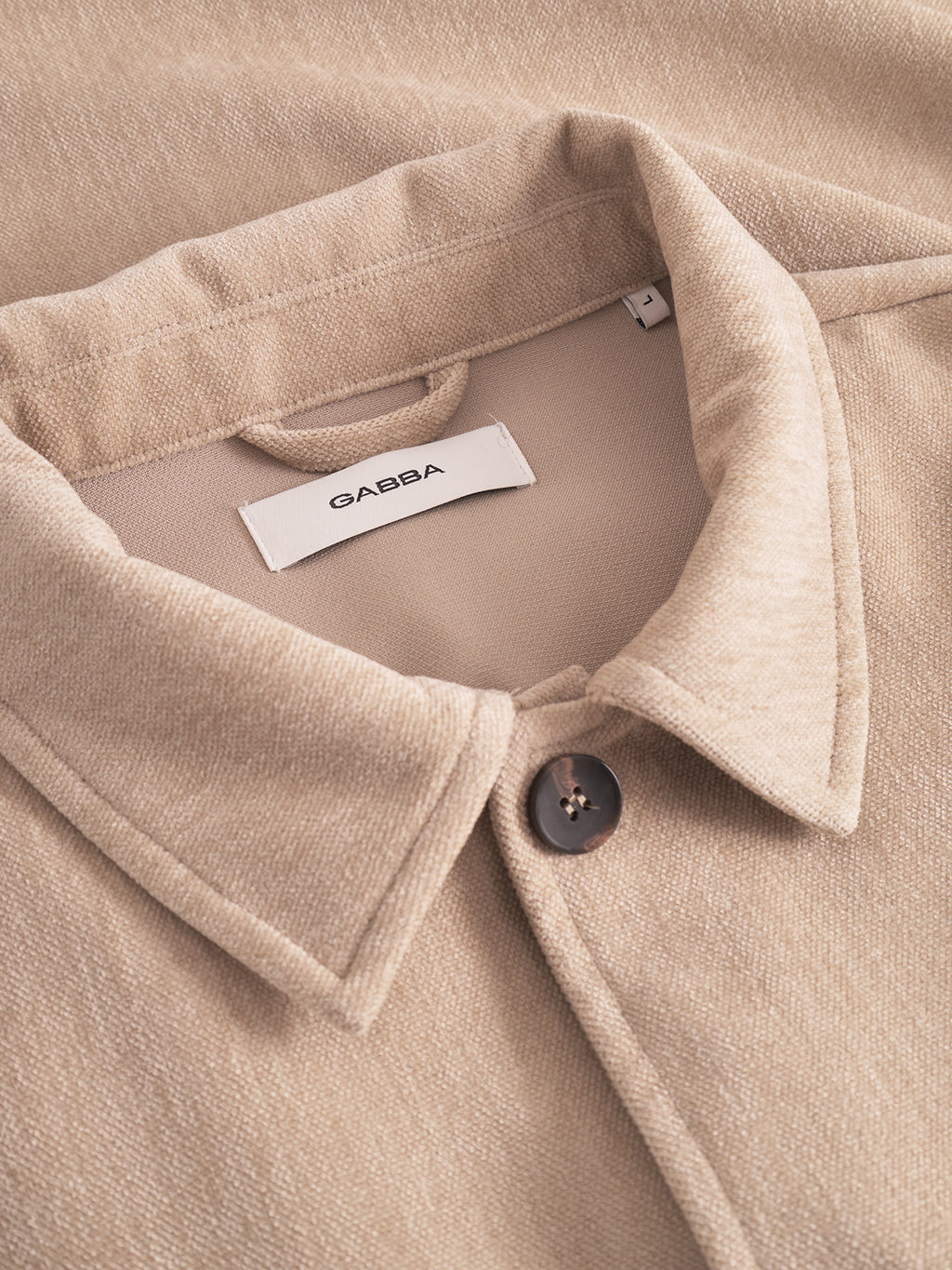 Close-up of Gabba’s Flap Velour - Lt. Sand overshirt, featuring a beige regular fit, black button, and soft textured fabric. A white Gabba label is visible inside the collar.