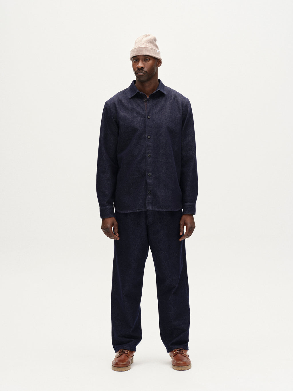 A man stands against a white background wearing the Gabba Sven Reg Chev dark navy workwear shirt and matching loose-fitting pants, paired with a beige beanie and brown shoes. He looks slightly to the side with a neutral expression.