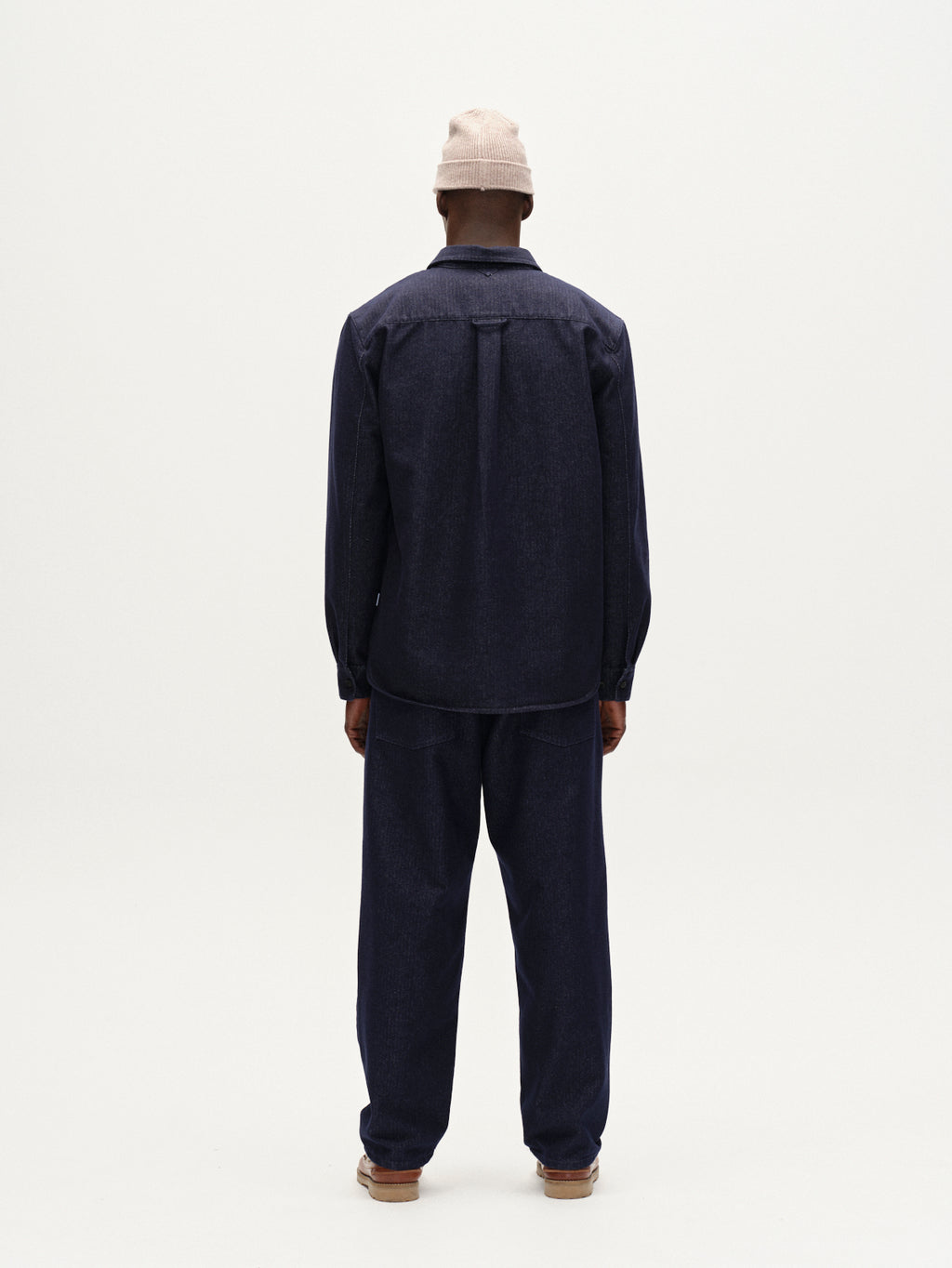 A person stands facing away from the camera, wearing the Gabba Sven Reg Chev - Dark Navy workwear shirt and pants, a light beige beanie, and brown shoes against a plain white background.