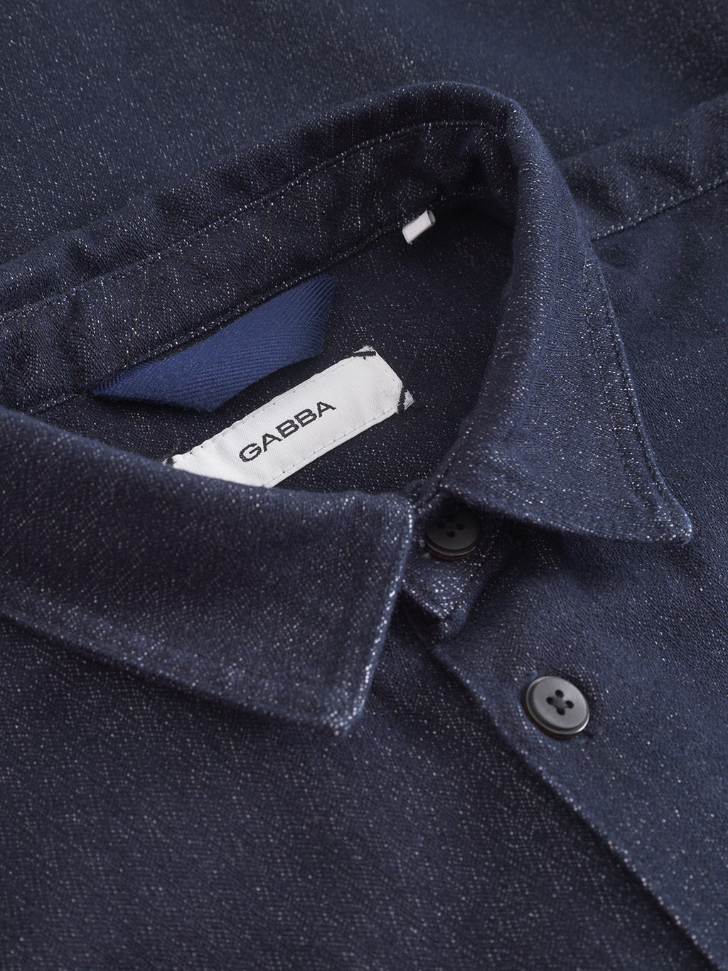 Close-up of the Sven Reg Chev - Dark Navy shirt by Gabba, showing black buttons and a Gabba fabric tag stitched inside the collar. The dark navy material displays a subtle, textured sheen with a herringbone pattern.