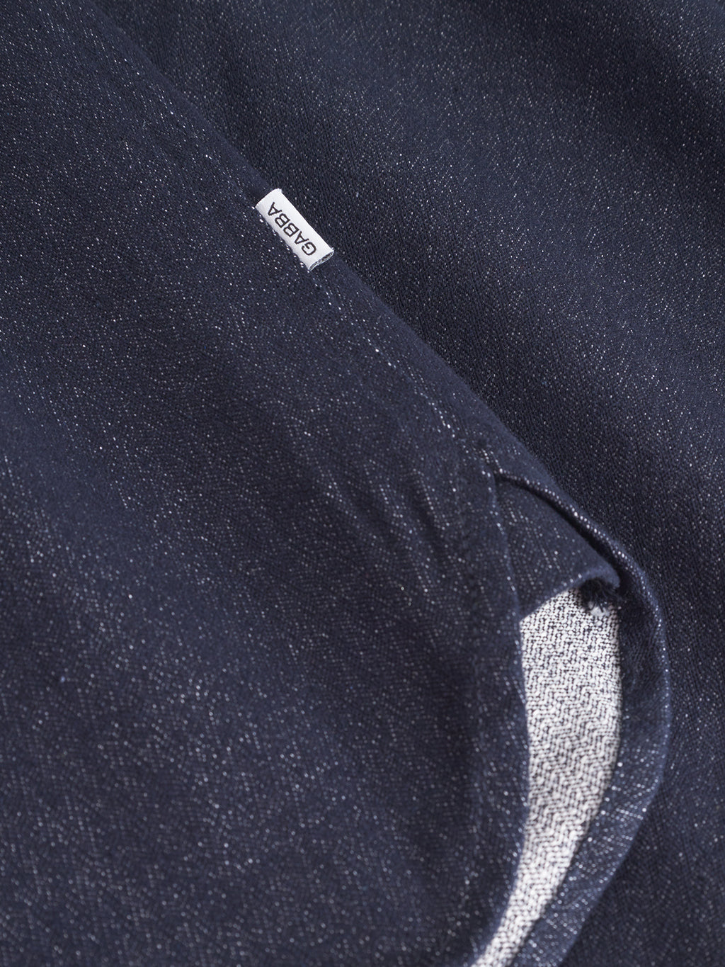 Close-up of Sven Reg Chev - Dark Navy denim shirt fabric by Gabba, showing a small white brand label and a folded edge that reveals the lighter, textured underside.