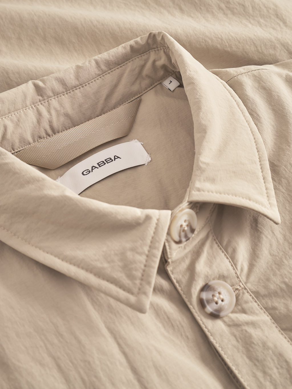 Close-up of the Gabba Nara Goz - Lt. Sand overshirt in beige, featuring white buttons and a collar tag with the brand name. The fabric is smooth, lightly textured, and padded—perfect as a light jacket.