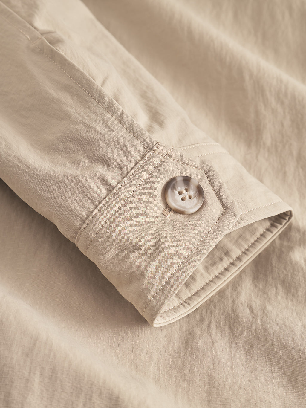 Close-up of the Gabba Nara Goz - Lt. Sand overshirt’s cuff, showing detailed stitching and a light button on padded beige fabric, laid flat atop matching material.