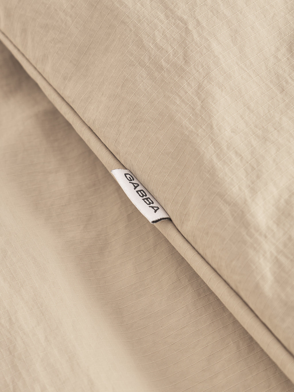 A close-up of Nara Goz - Lt. Sand fabric by Gabba shows its beige woven texture and a white Gabba tag, ideal for a slightly padded light jacket or regular fit overshirt.