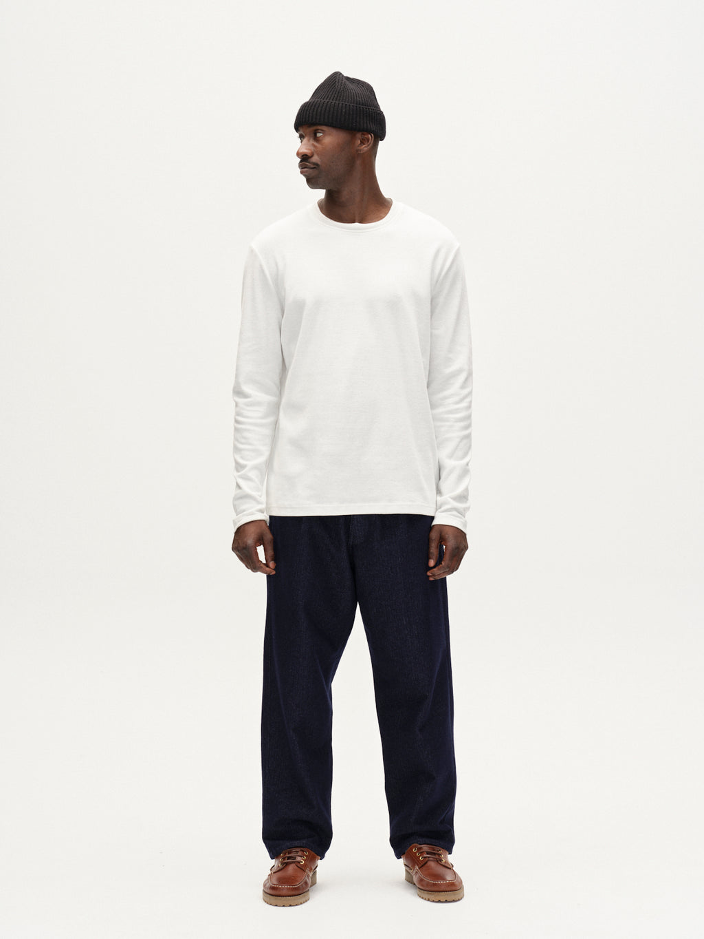 A man in a Gabba Dune Brush LS - Off White long-sleeve tee, black beanie, loose dark blue pants, and brown shoes stands slightly turned left against a plain white background.