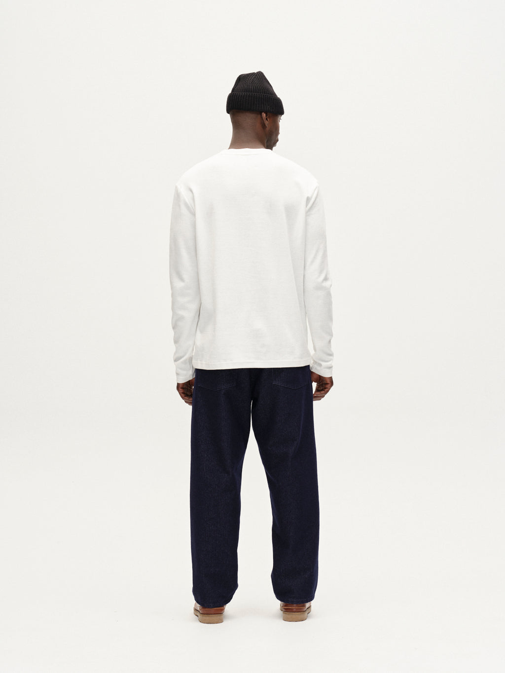 A person stands facing away, wearing the Gabba Dune Brush LS - Off White long-sleeve tee, paired with dark blue pants, brown shoes, and a black beanie against a plain white background.