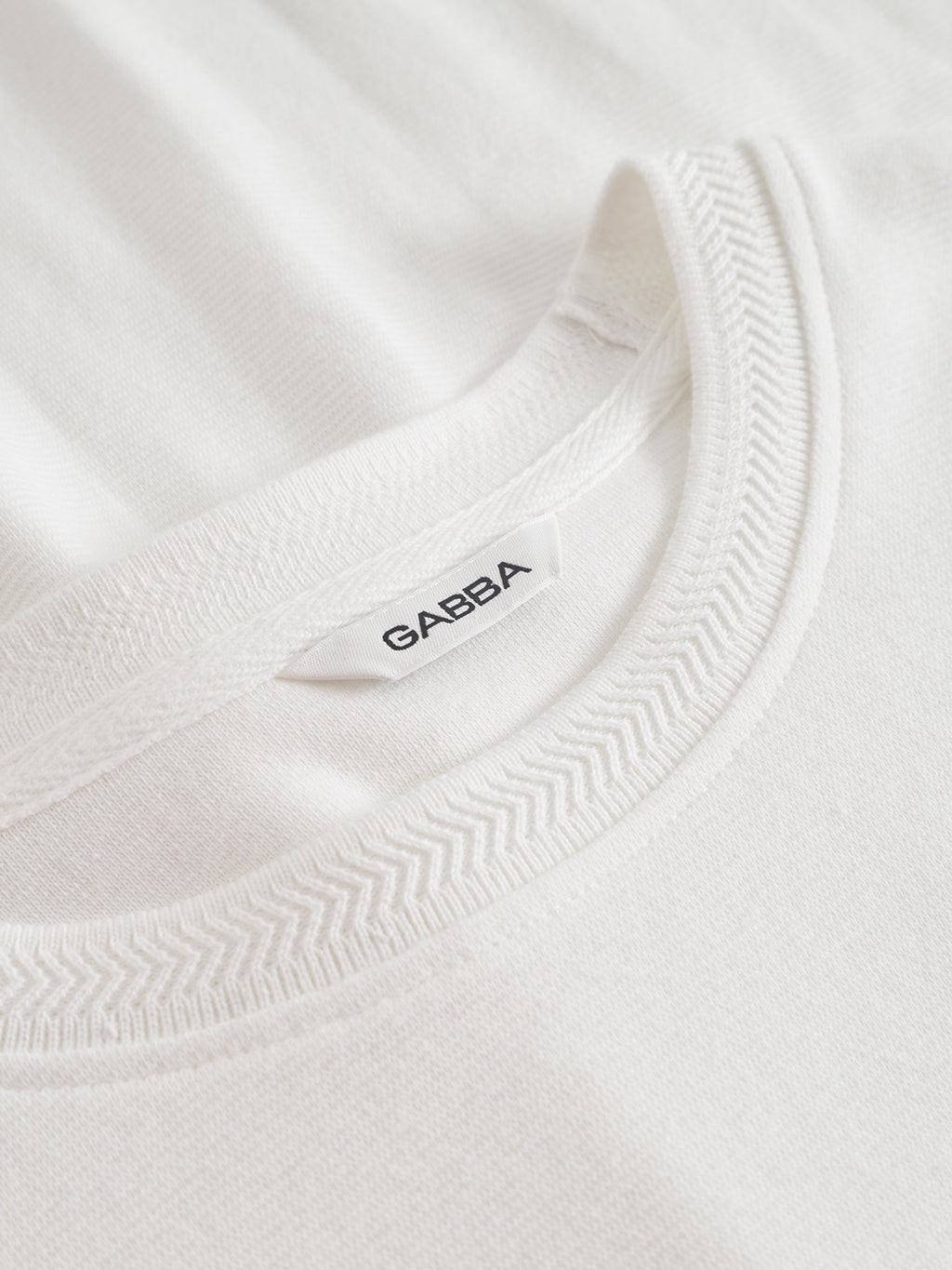 Close-up of the collar of the Gabba Dune Brush LS in Off White, featuring a white tag with the Gabba logo in black. The stitching and textured fabric surface are clearly visible.