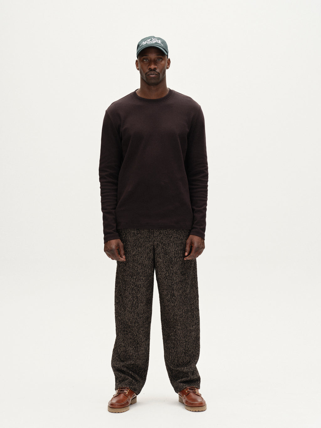 A man stands against a plain white background, wearing the Gabba Dune Brush LS - Black with loose patterned trousers, brown shoes, and a green baseball cap. He faces forward, arms relaxed at his sides.