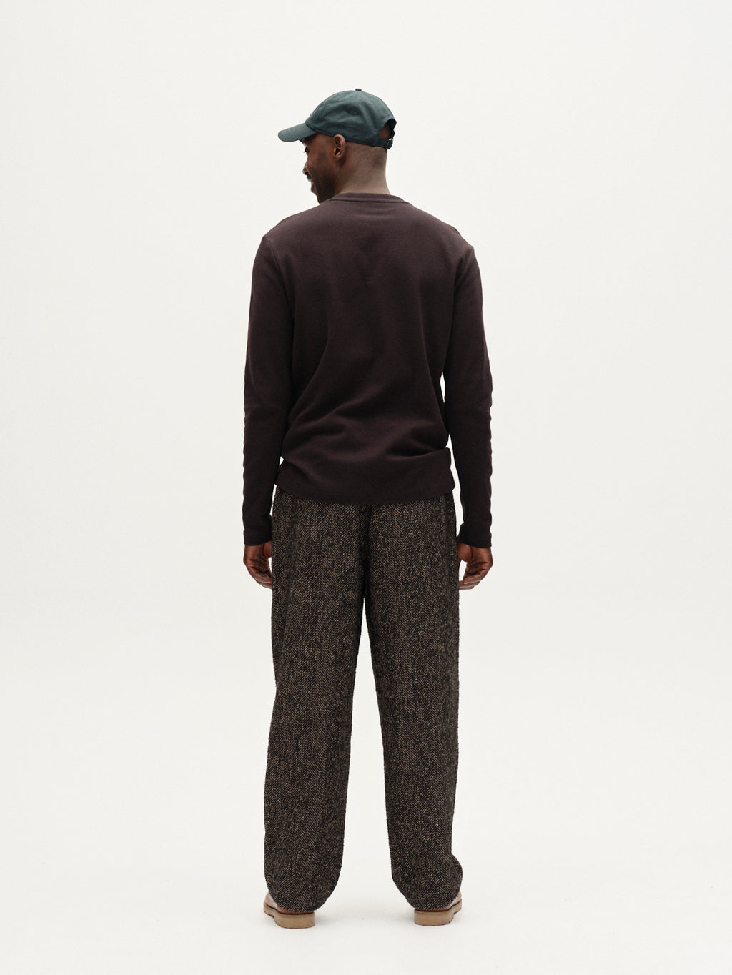 A man stands facing away from the camera, wearing Gabba’s Dune Brush LS in black, loose textured gray pants, brown shoes, and a dark green cap—an effortlessly modern look—set against a plain white background.