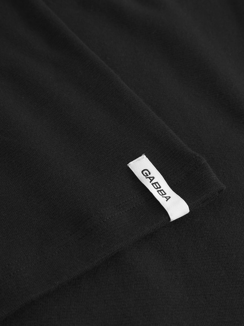 A close-up of the Dune Brush LS - Black by Gabba, a modern long-sleeved tee, shows a small white tag with GABBA in black capital letters sewn into the seam.