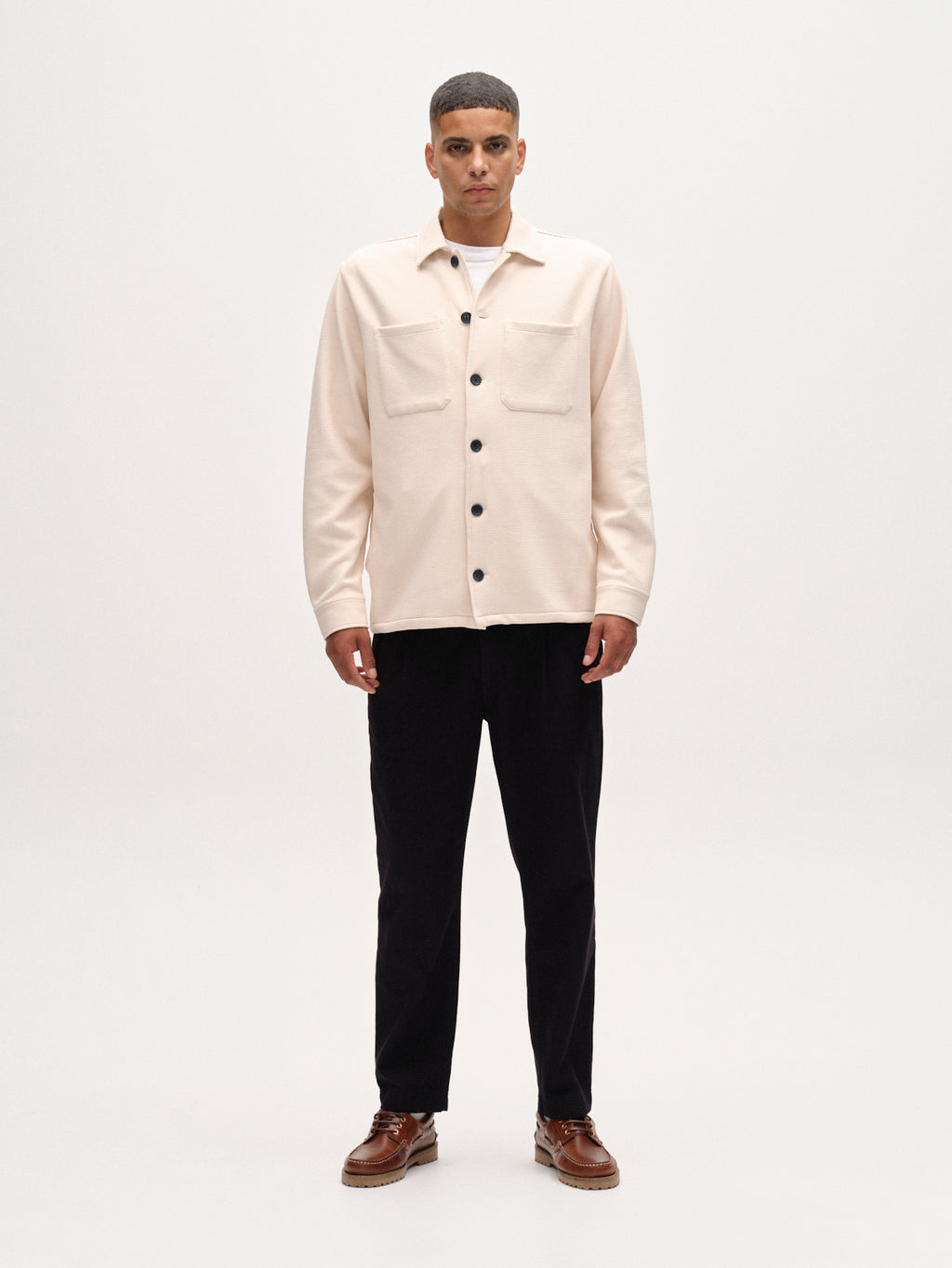 A man stands facing forward against a plain white background, wearing the Gabba Como Waff overshirt in White Almond, paired with black pants and brown shoes.