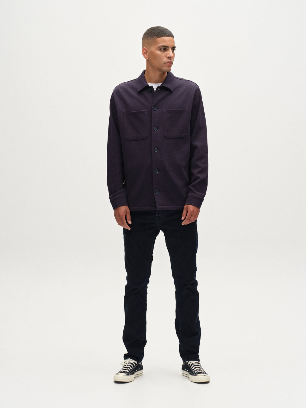A man stands against a white background, wearing the Gabba Como Waff overshirt in navy with large chest pockets, paired with dark jeans and black-and-white sneakers. He looks slightly left with his hands at his sides.