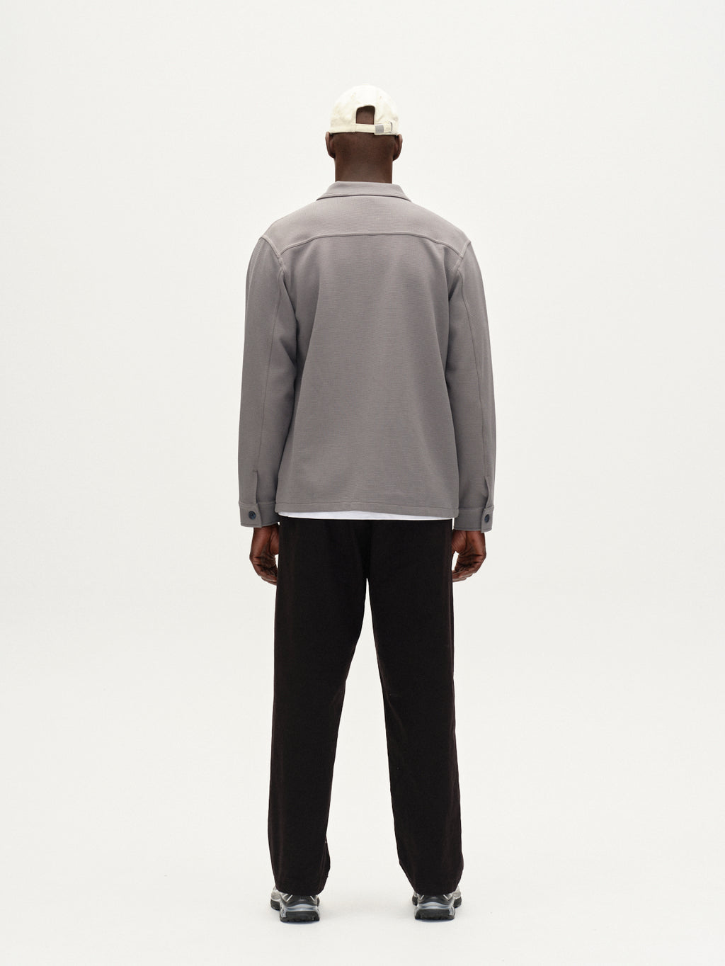 A person faces away from the camera, wearing Gabbas Como Waff overshirt in Dust Sage Green over a light gray jacket, paired with black pants, black sneakers, and a white baseball cap against a plain light background.