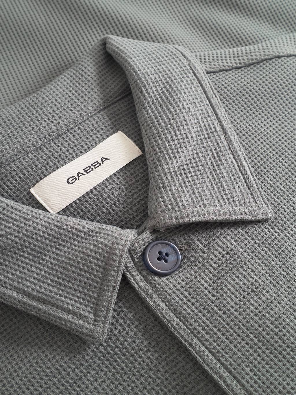 Close-up of the Gabba Como Waff collar in dust sage green, featuring a single blue button and inner white brand label—a versatile piece that layers perfectly beneath a waffle-knit overshirt.